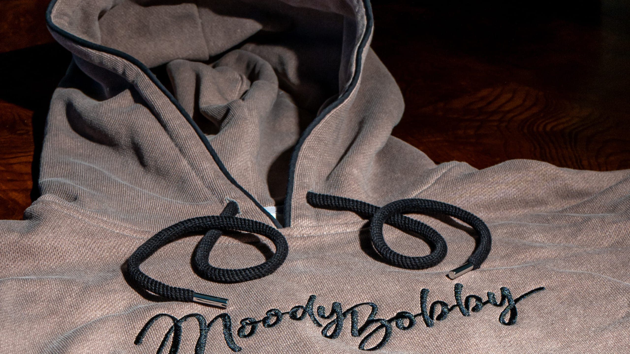 Close view of the embroidered Moody Bobby script stitched across the chest of an oatmeal hoodie, lit low and warm
