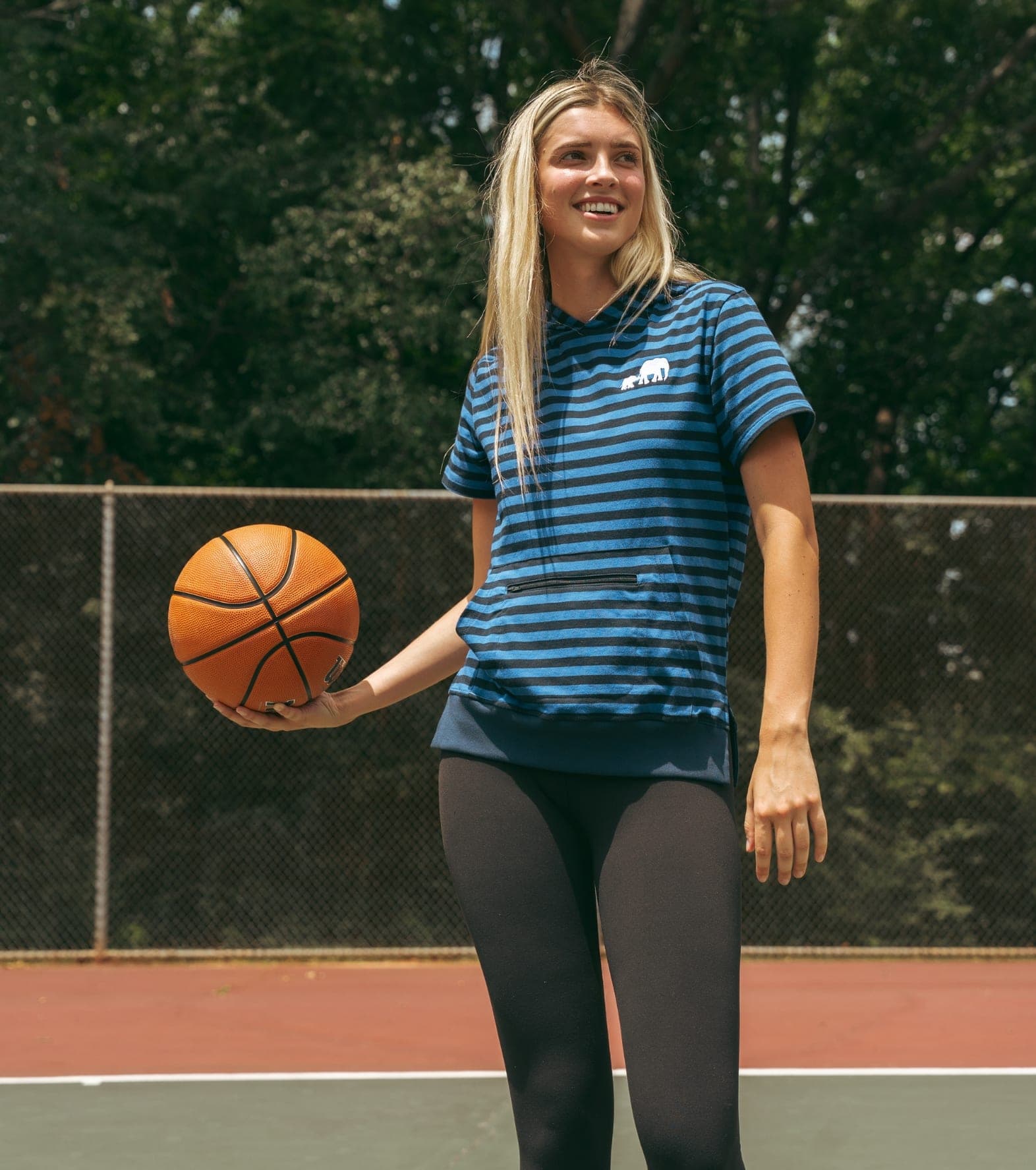 Woman in a blue striped tee standing on an outdoor court, holding a basketball at her side